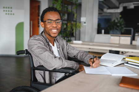 Happy young black man with disability using a wheelchair is taking notes while studying in a bright...