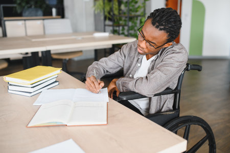 African American student in a focusing wheelchair on writing notes at a desk, surrounded by books in...