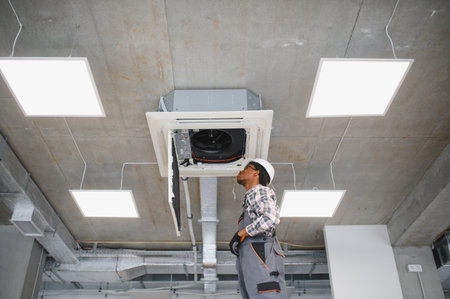 African american technician wearing safety gear works on repairing a ceiling air conditioning unit...