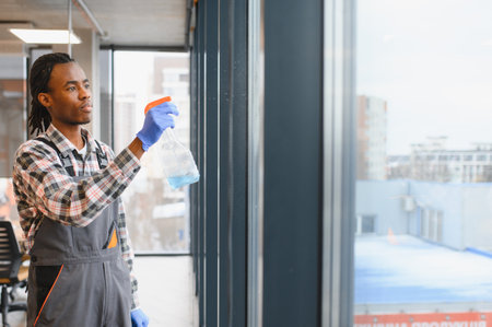 Professional worker in overalls cleaning windows of a modern office building, using a spray bottle and wearing protective gloves for safetyの写真素材