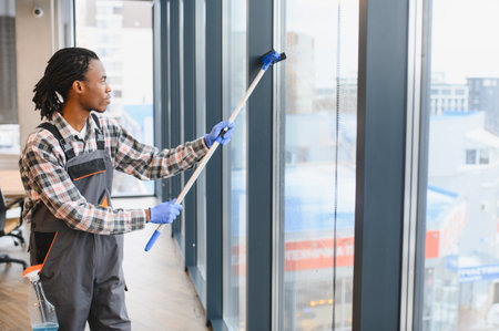 Young man cleaning windows in an office building, using a squeegee and cleaning liquid to ensure a spotless, hygienic environmentの写真素材
