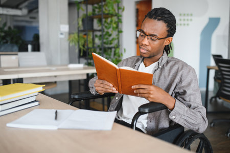 Young black man with disability using a wheelchair, concentrating on his studies while reading a...