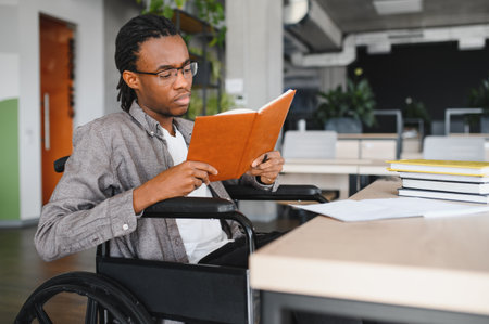 Young black man with disability using wheelchair, concentrating on his studies, reading textbook in a bright, contemporary office spaceの写真素材