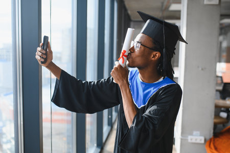Happy african american graduate student kissing diploma and taking selfie with smartphone near...