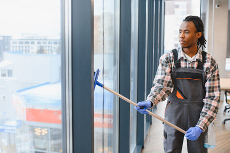 African american worker efficiently cleaning large office windows with a squeegee, wearing protective gloves and overalls, ensuring clear visibilityの写真素材