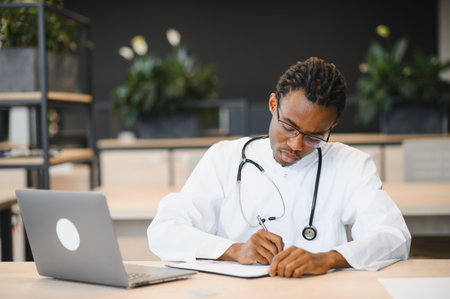 Focused young male doctor writing notes at a desk with a laptop, surrounded by greenery, showcasing a professional healthcare environmentの写真素材