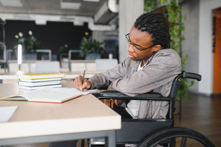 Young African American student in a wheelchair focused on writing notes at a desk in a modern, well lit study spaceの写真素材
