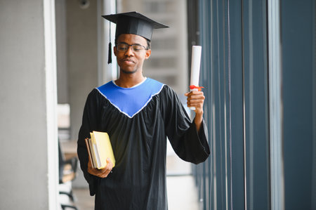 Young african american student wearing graduation gown is holding his diploma and books in...