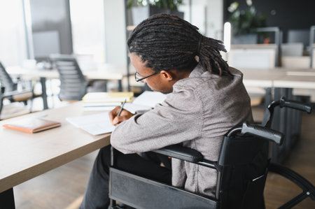 Young black man with disability using wheelchair studying and taking notes in bright coworking spaceの写真素材
