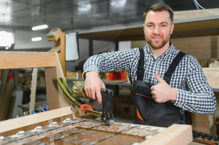 Carpenter assembling wooden sofa frame using pneumatic staple gun and showing thumbs up gesture in...