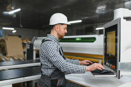 Skilled carpenter wearing safety helmet operating cnc machinery in a modern furniture factory,...