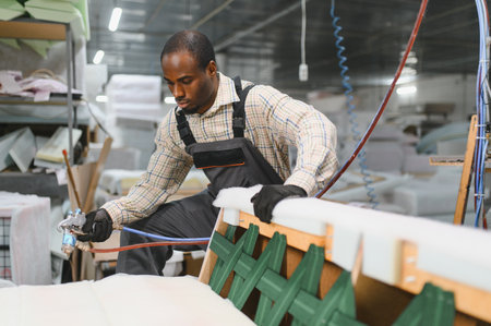 Concentrated african american craftsman using glue spray gun on sofa frame in furniture factoryの写真素材