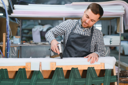 Carpenter operating a pneumatic staple gun while upholstering a sofa in a bustling furniture factory, showcasing skilled craftsmanshipの写真素材