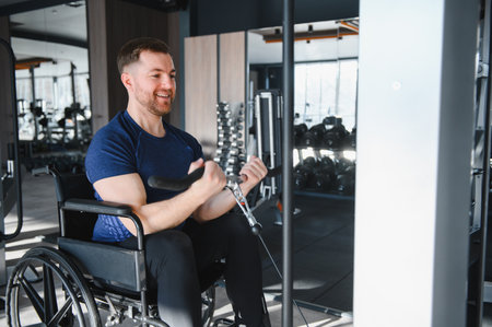 Smiling man in wheelchair exercising his biceps with weights in gym, demonstrating sports...