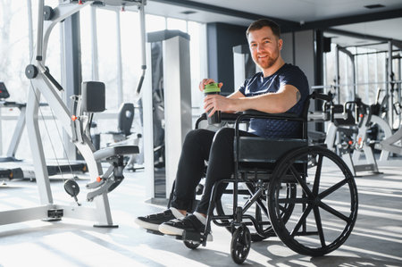 Smiling sportsman drinking from a green water bottle, sitting in a wheelchair, resting after exercising in a modern gymの写真素材