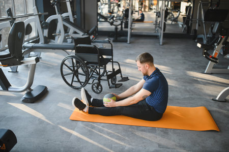 Person using medicine ball while training on orange mat in gym, with wheelchair nearby, promoting rehabilitation and inclusive fitnessの写真素材