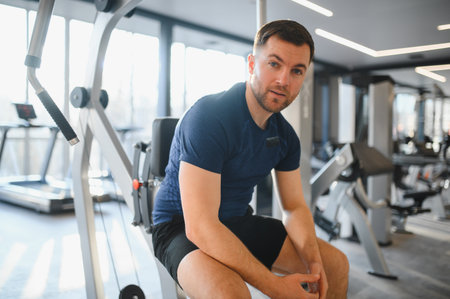 Young man sitting and resting on fitness equipment, recovering after physical activity in modern gym