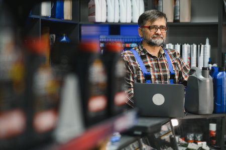 Auto parts store salesman wearing blue dungarees using laptop with shelves full of motor oil and car...
