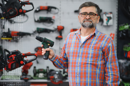 Senior salesman holding a cordless drill in a hardware store, power tools displayed on shelves in...