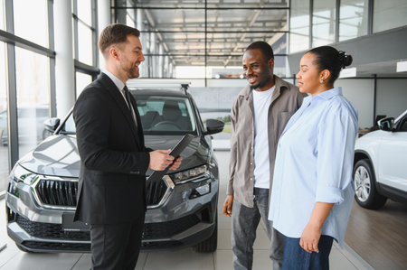 Smiling car salesman holding clipboard presenting new car to african american couple in modern...