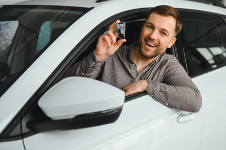 Enthusiastic man sitting in a car, holding a key with a smile, showcasing excitement for his new vehicle in a modern showroomの写真素材