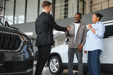 Happy african american couple receiving car keys from salesman in a car dealership, buying a new vehicleの写真素材