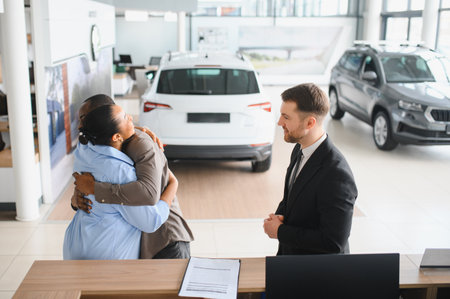 Couple embracing joyfully after purchasing a new car at the dealership, with a salesman observing their excitement and happinessの写真素材