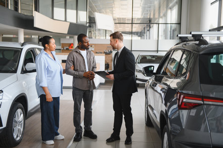 Car salesman assisting an African American couple while showcasing vehicles in a bustling dealership, discussing purchase options for their new carの写真素材