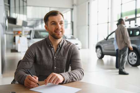 Happy customer signing contract after choosing new car in dealership showroom, salesman assisting customer in backgroundの写真素材