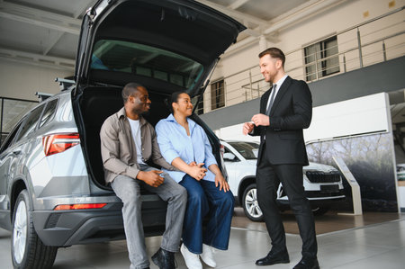 Happy couple sitting in open trunk of new car while salesman explains features in modern dealership