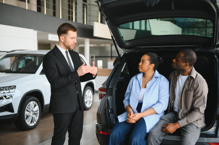 Car salesman explaining trunk features to African American couple sitting in open trunk, considering...