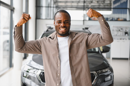 Excited african american man flexing biceps after purchasing vehicle in car dealership, celebrating new purchaseの写真素材