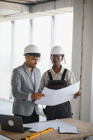 Architect and builder wearing hardhats reviewing blueprint together on construction site, discussing building plans and project progressの写真素材