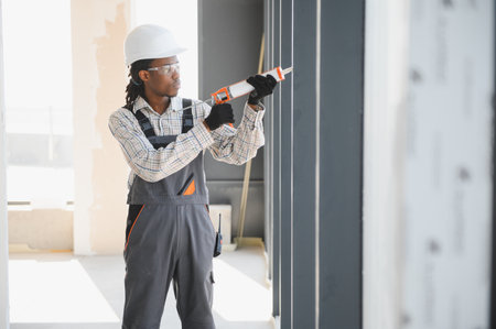 African american construction worker applies sealant to a window frame on a construction site,...