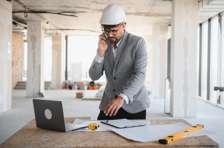 Arab architect wearing suit and hardhat managing construction site, talking on phone and checking...