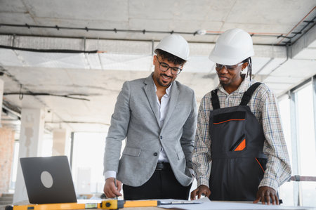 Architect and builder wearing safety helmets are examining blueprints on a construction site, using...