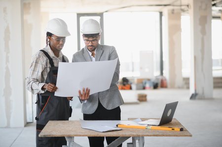 Architect and builder wearing hardhats and examining blueprints on a construction site, discussing...