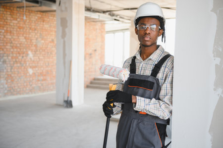 Young african american construction worker holding paint roller, wearing safety glasses and hardhat,...