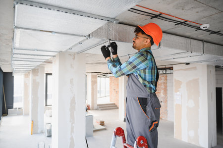 Electrician working on a ventilation system while on a ladder in an unfinished building, ensuring...