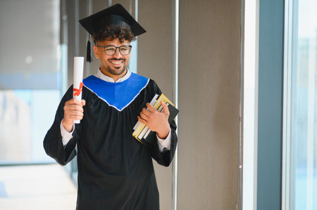 Smiling arab student wearing a graduation gown, proudly holding a diploma and books while...