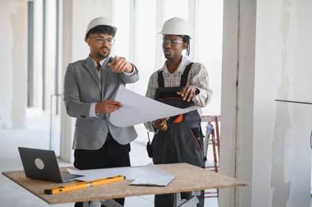 Architect and builder wearing hardhats reviewing blueprints during a construction meeting,...