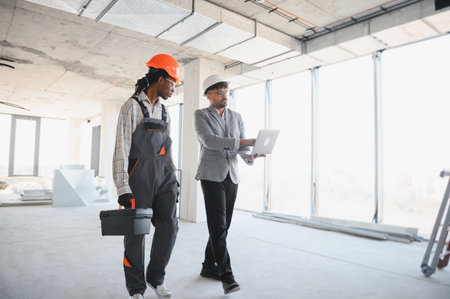 Architect showing building plans on laptop to builder walking inside building under construction