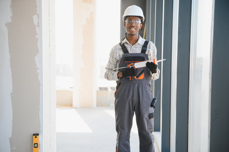 Smiling african american construction worker holding silicone sealant gun while standing near window...
