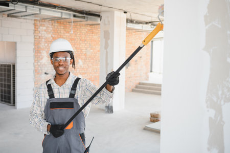 Smiling african american construction worker painting a wall using a telescopic paint roller on a...