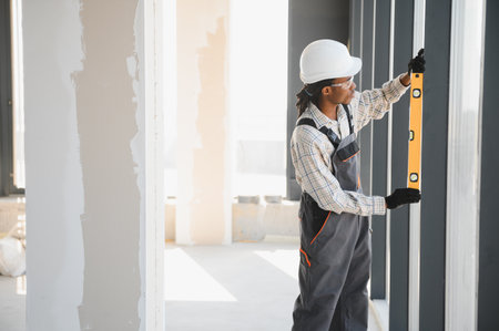 African american builder using spirit level while working on window installation in building under...