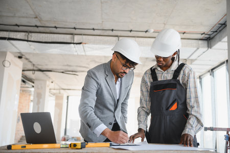Architect and construction worker wearing hardhats reviewing blueprints in a building under...