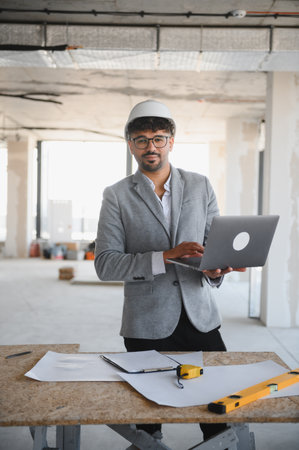 Arab architect wearing hardhat using laptop at construction site, with blueprint and level on table