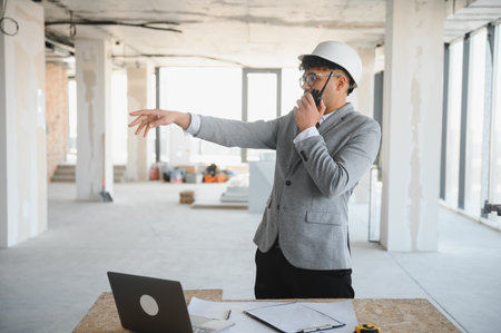 Arab architect wearing suit and hardhat using walkie talkie and pointing at something during...