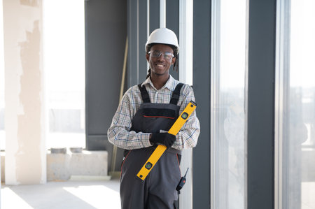 African american construction worker wearing safety helmet and work overalls holding a spirit level...