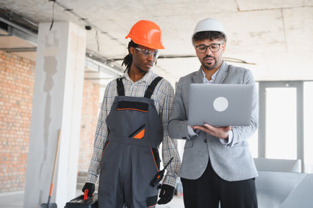 Architect showing building plans on a laptop to a builder during a construction site inspection